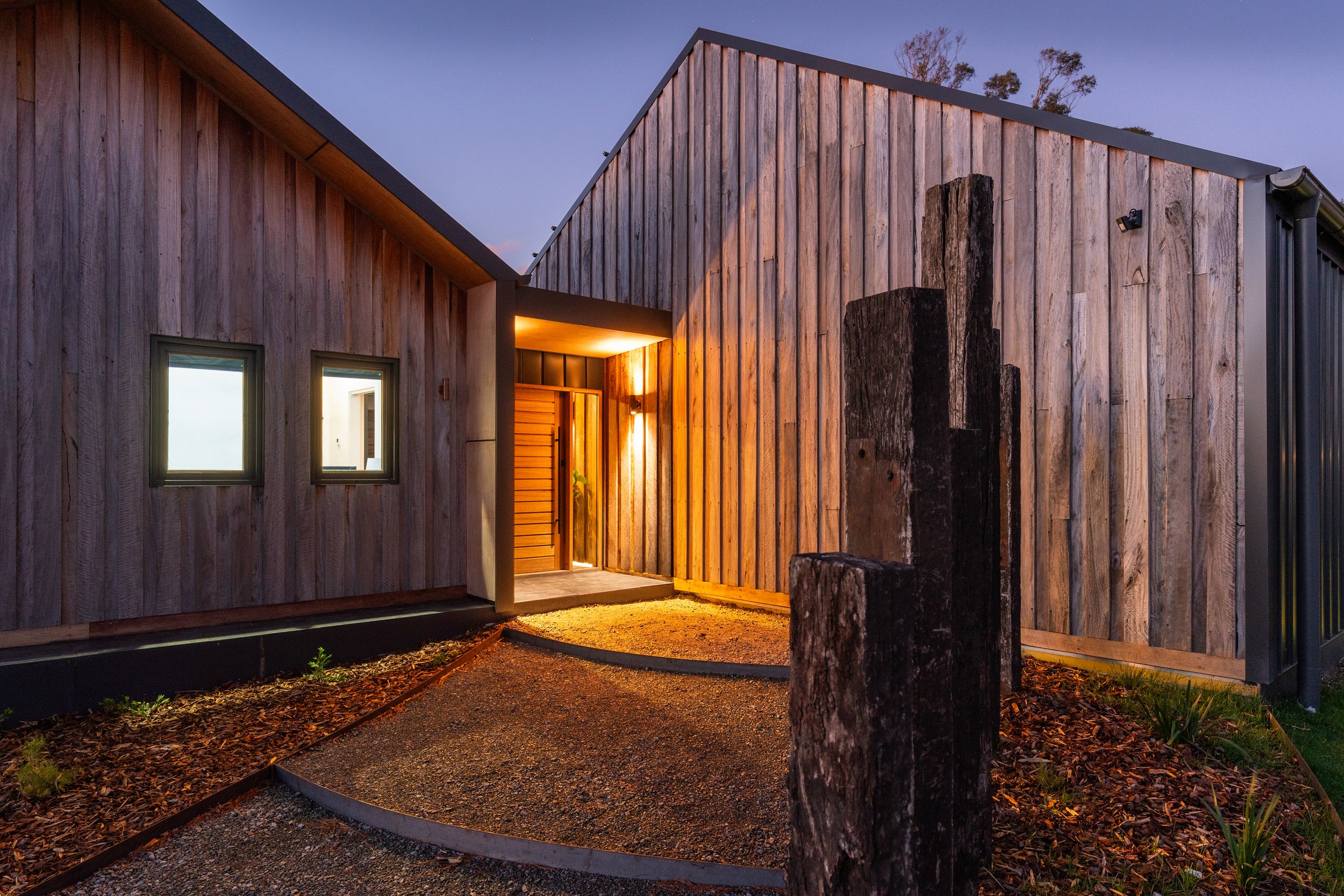 Modern house with wooden exterior during dusk, lit warmly at the entrance, with a gravel pathway and wooden posts in front.
