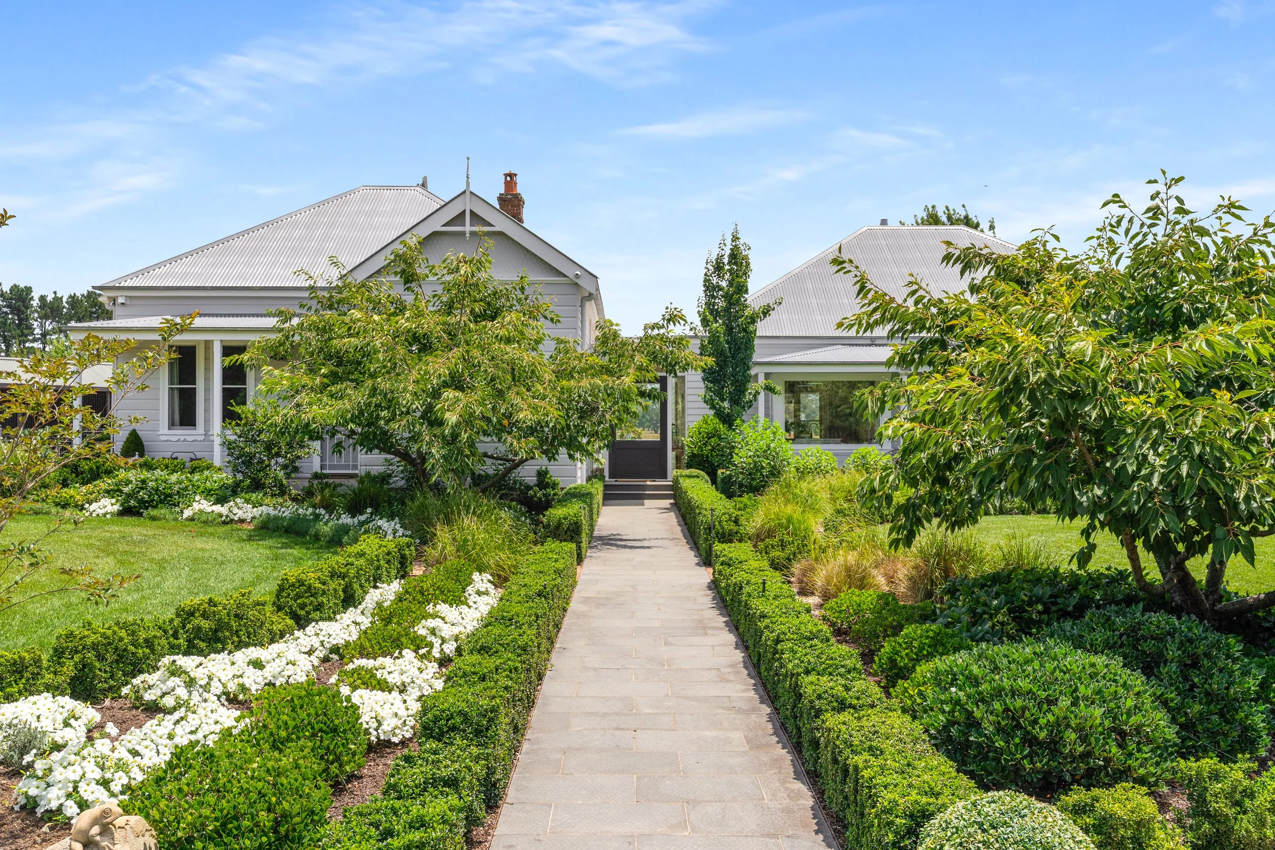 A paved walkway leading to a white house with a metal roof, surrounded by lush green trees, bushes, and flowering plants on a sunny day.