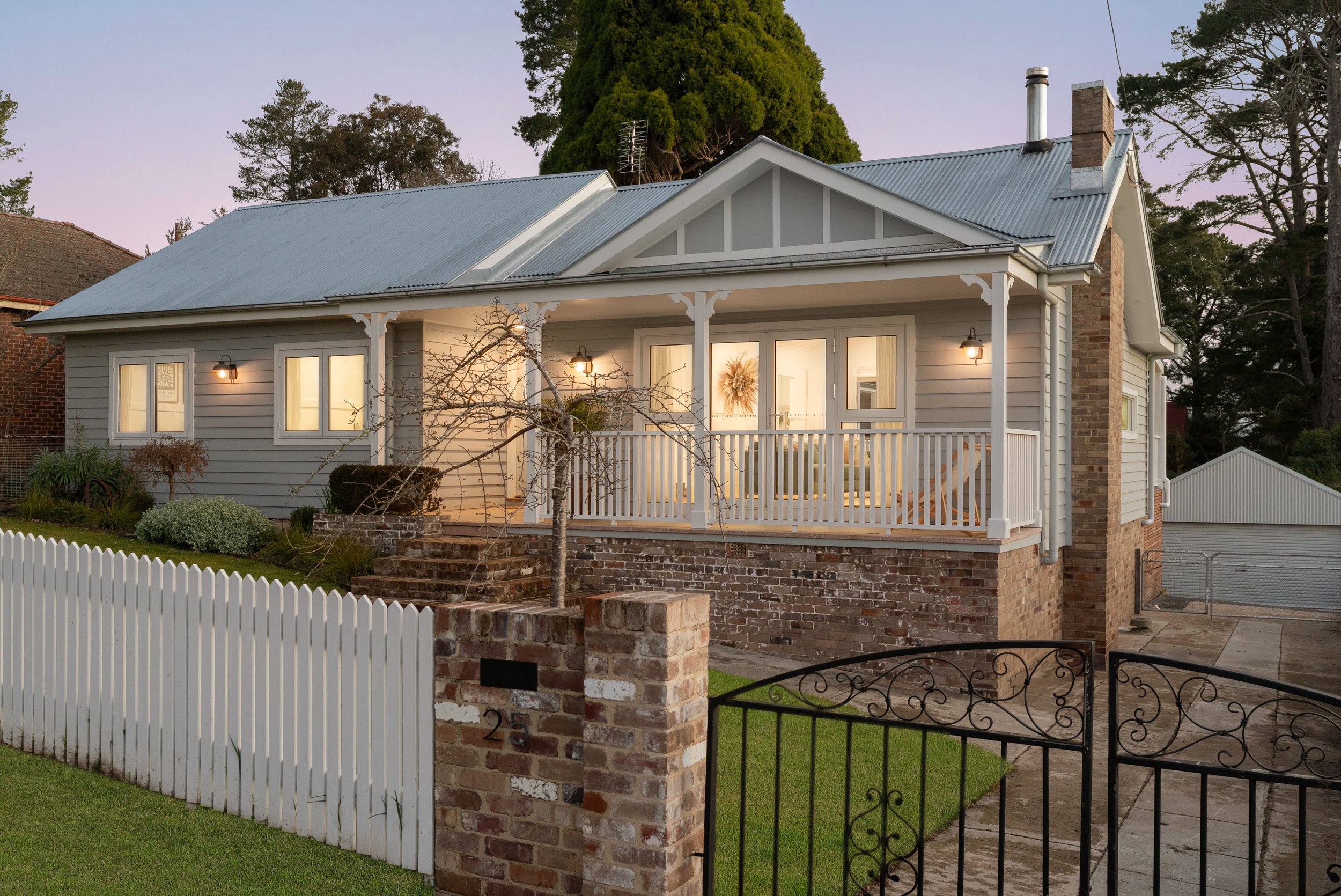 A cozy, two-story house with a white wooden exterior, metal roof, brick foundation, and front porch illuminated with warm lighting, surrounded by a white picket fence and a decorative black gate, during dusk.