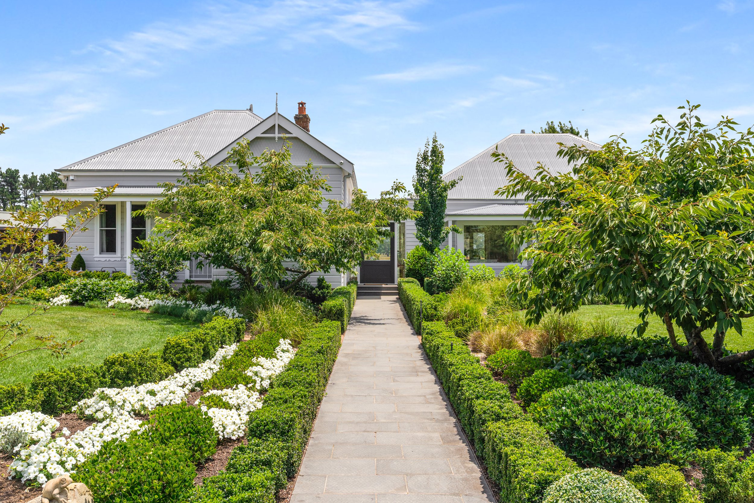 A paved walkway leading to white houses with gray roofs, surrounded by lush green trees and shrubs, under a clear blue sky.