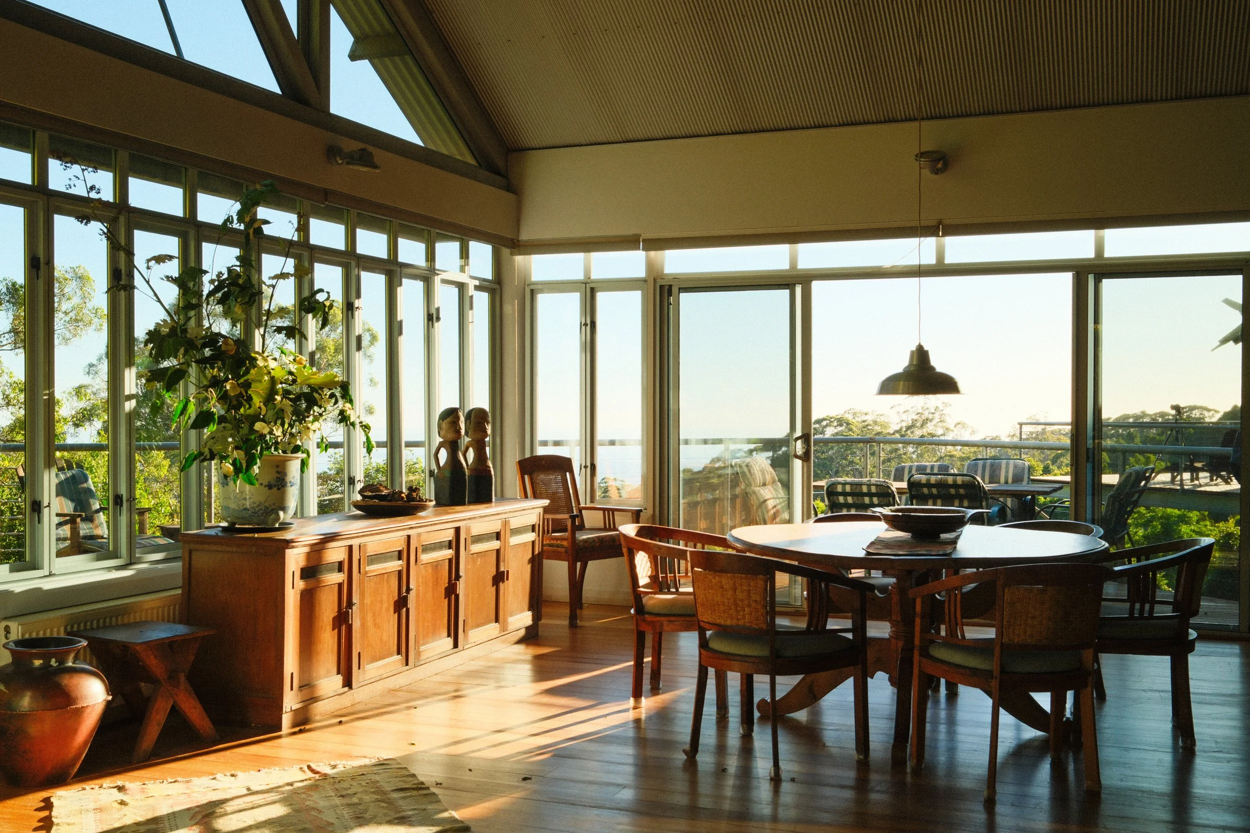 Sunlit dining area with wooden furniture, large windows with a view of greenery, potted plants, decorative pieces, and outdoor seating on a balcony.