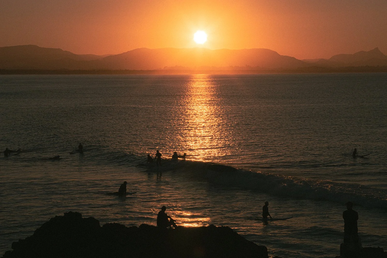 Silhouettes of people surfing and standing in the ocean during a sunset, with mountains in the background and the sun reflecting on the water.