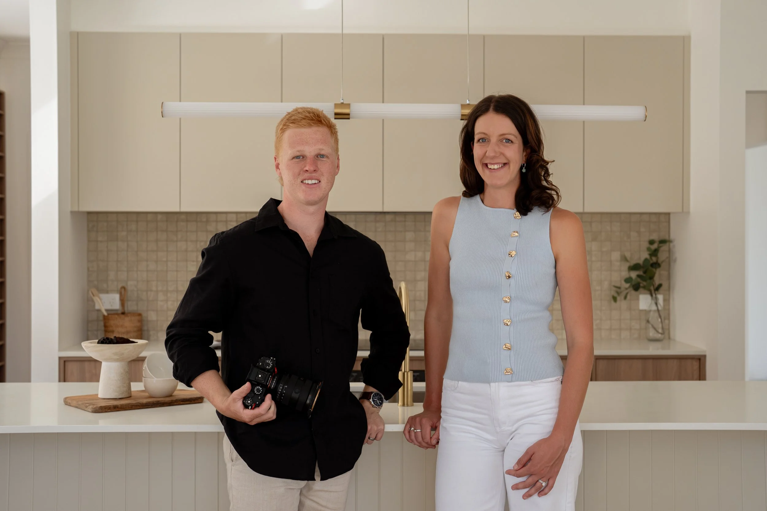 A man and woman standing in a modern kitchen, smiling, with the man holding a camera.