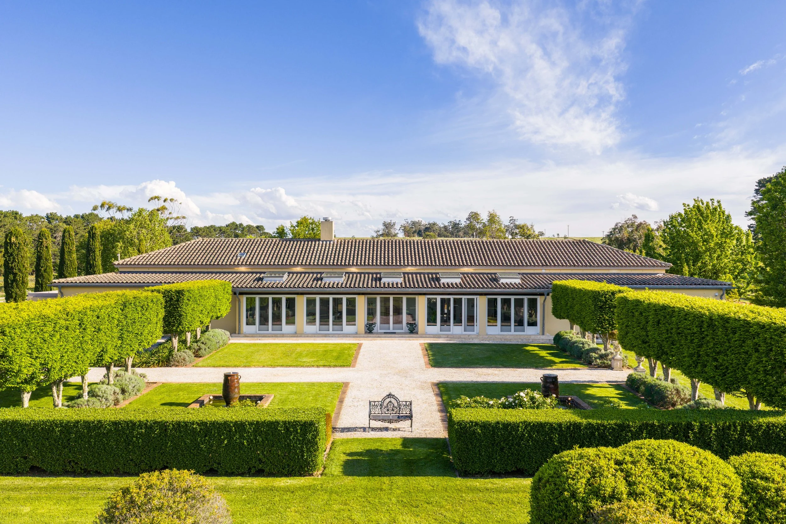 Large house with glass doors, surrounded by lush manicured gardens and tall trees under a blue sky with scattered clouds.