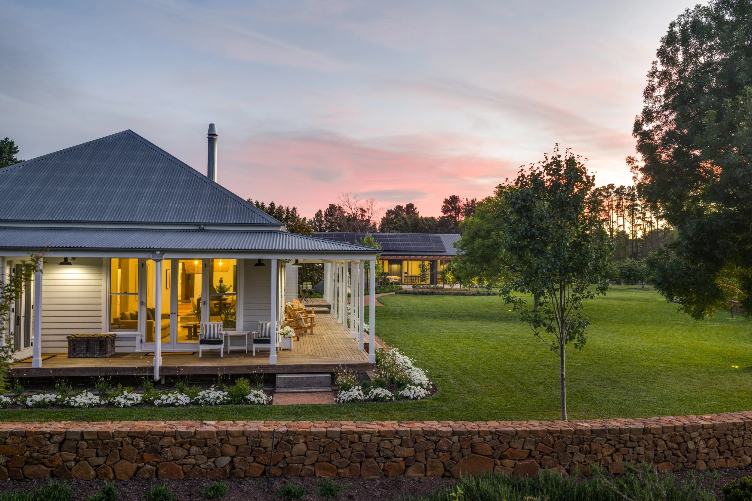 A house with a patio lit from inside, overlooking a well-manicured lawn with trees, under a sunset sky.