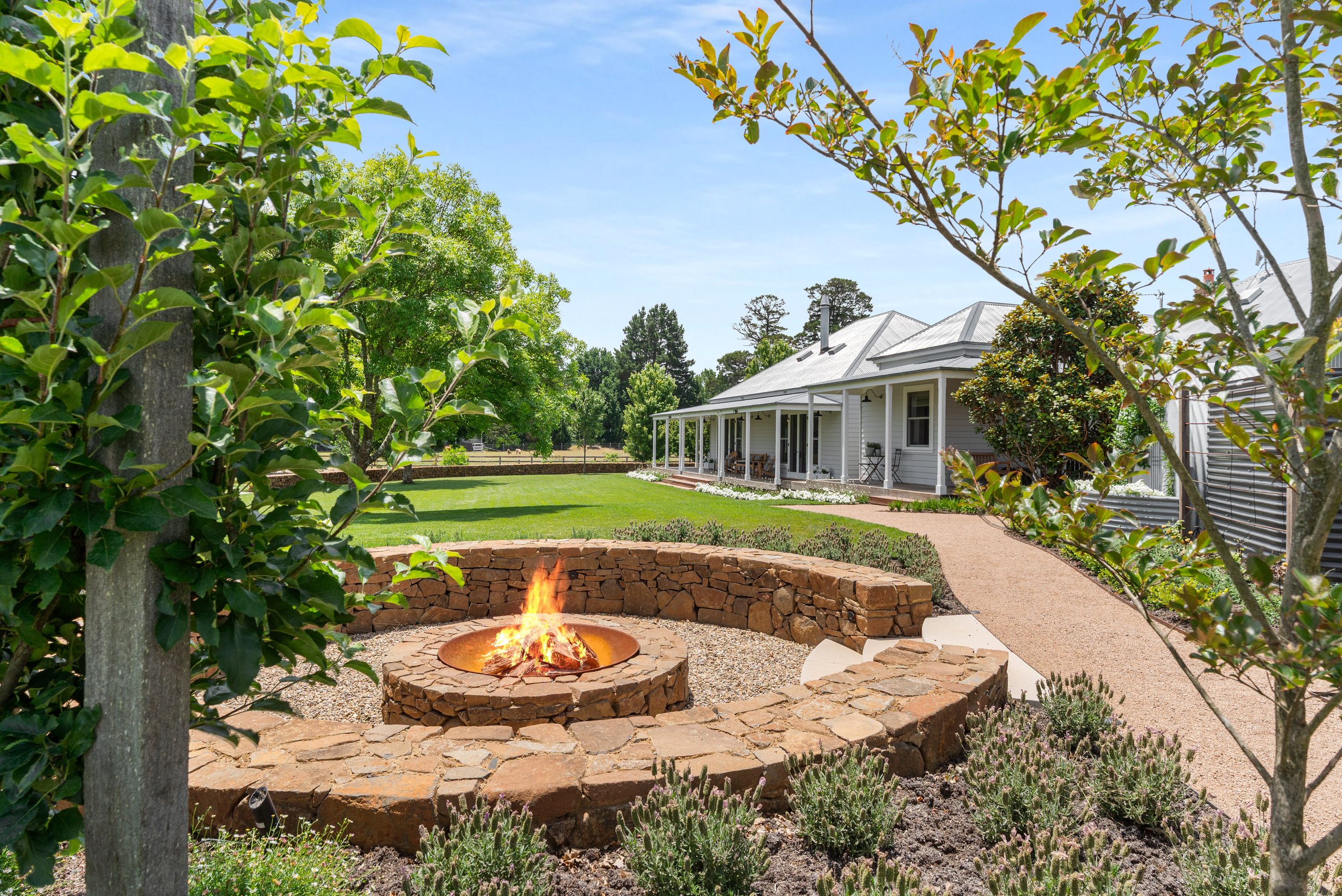 A backyard with a stone fire pit, green grass, trees, and a white house with a porch.