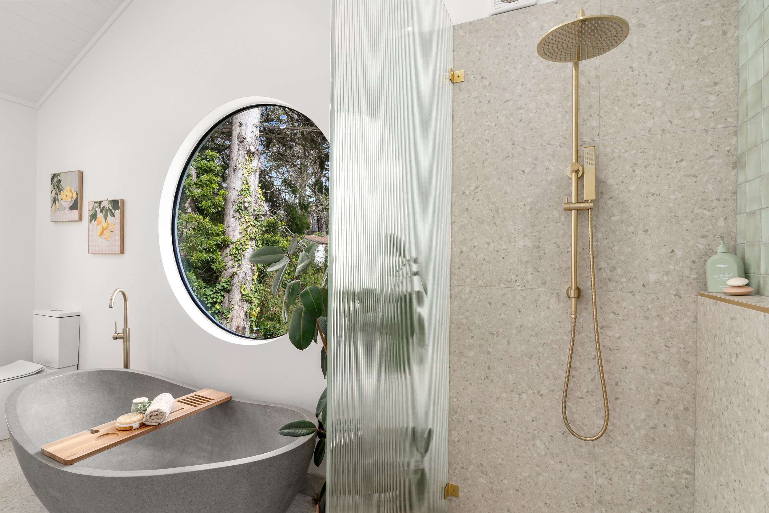 Modern bathroom with a freestanding stone bathtub, round window showing trees outside, and a walk-in shower with gold fixtures, beige textured wall, and minimal decor.