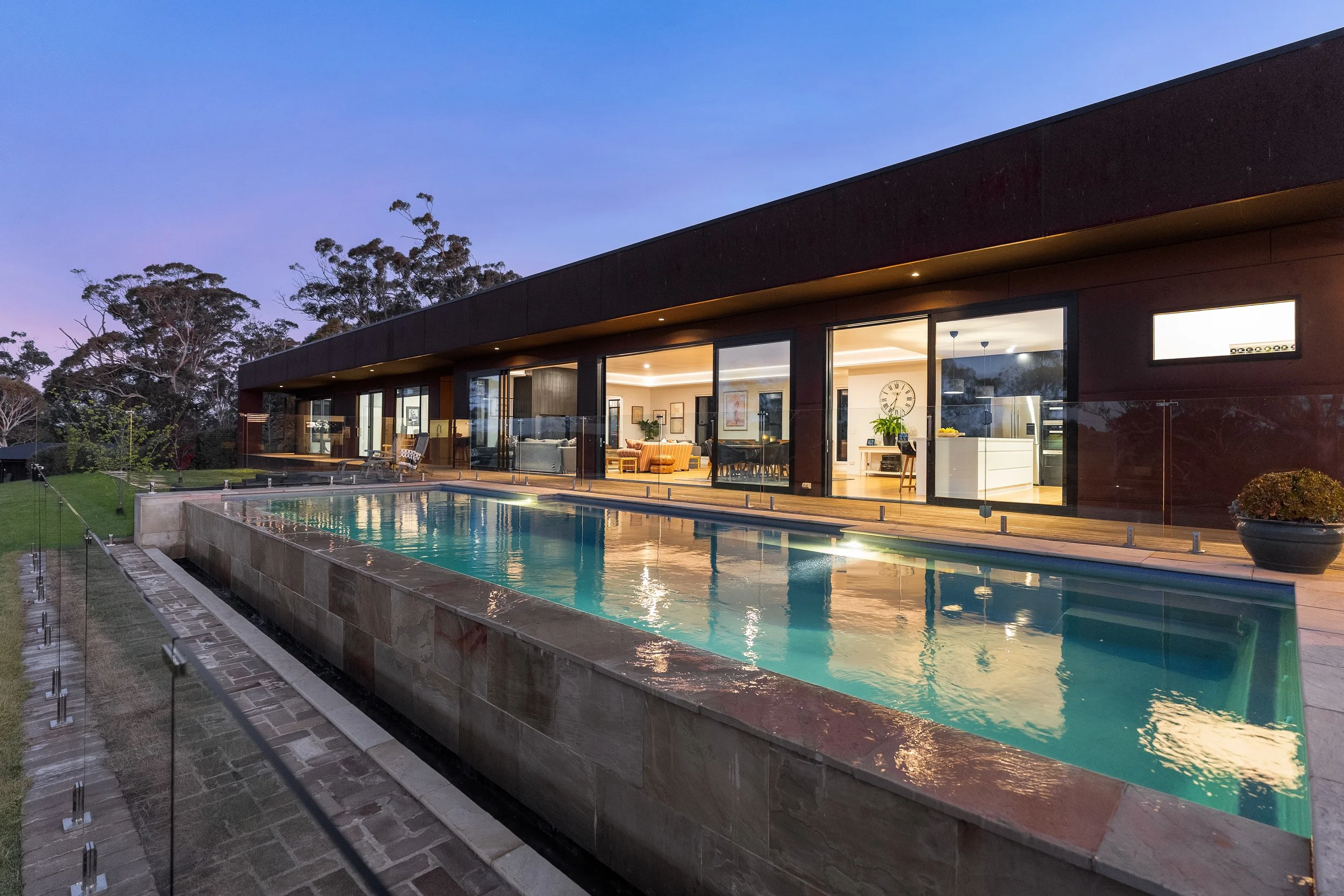 Modern house with large glass windows and a lit interior, adjacent to a rectangular swimming pool with a glass fence at dusk, surrounded by green grass and trees.