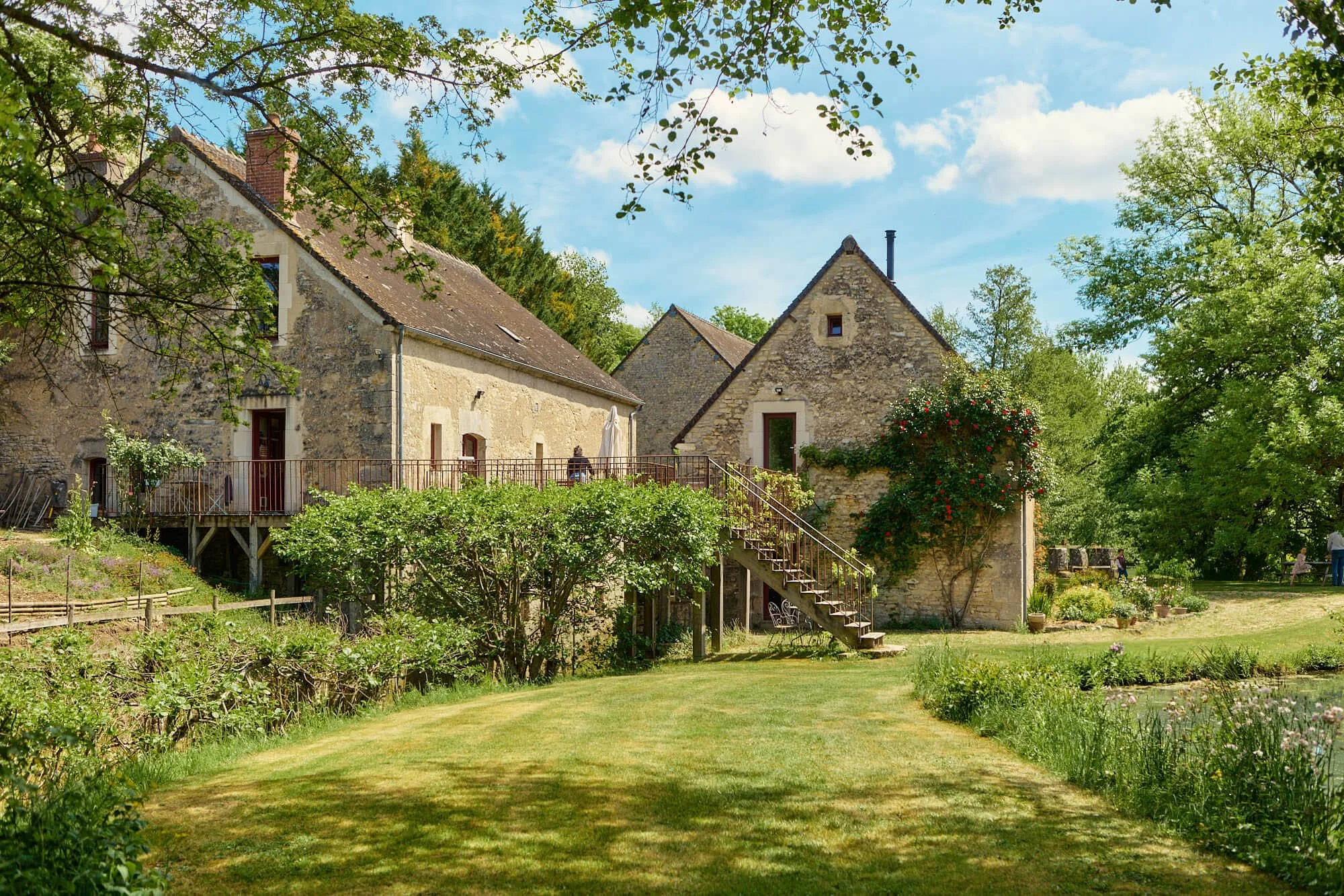 A stone house with multiple sections, surrounded by green trees and a well-maintained lawn. There is an outdoor staircase leading to a deck, and a person sitting at a table on the deck. The scene is sunny with a blue sky and scattered clouds.