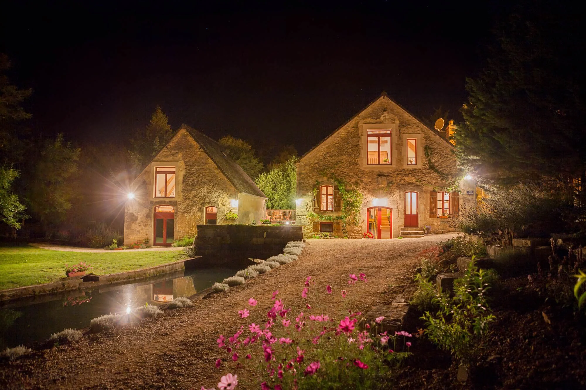 Exterior of the historic mill and gîte at night, with warm lights highlighting the buildings and creating a cosy, atmospheric scene.