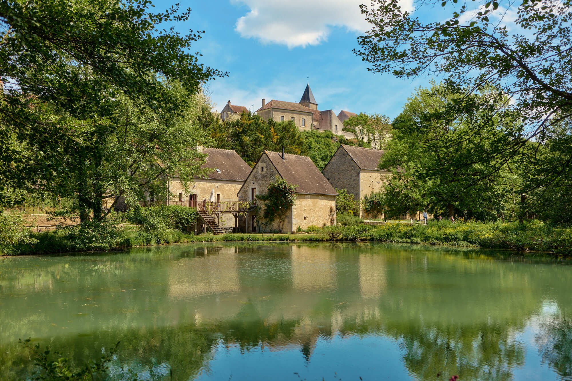 Panoramic view across the lake capturing the mill ensemble with the gîte at its centre, framed by the village of Rix rising on the hillside in the background.