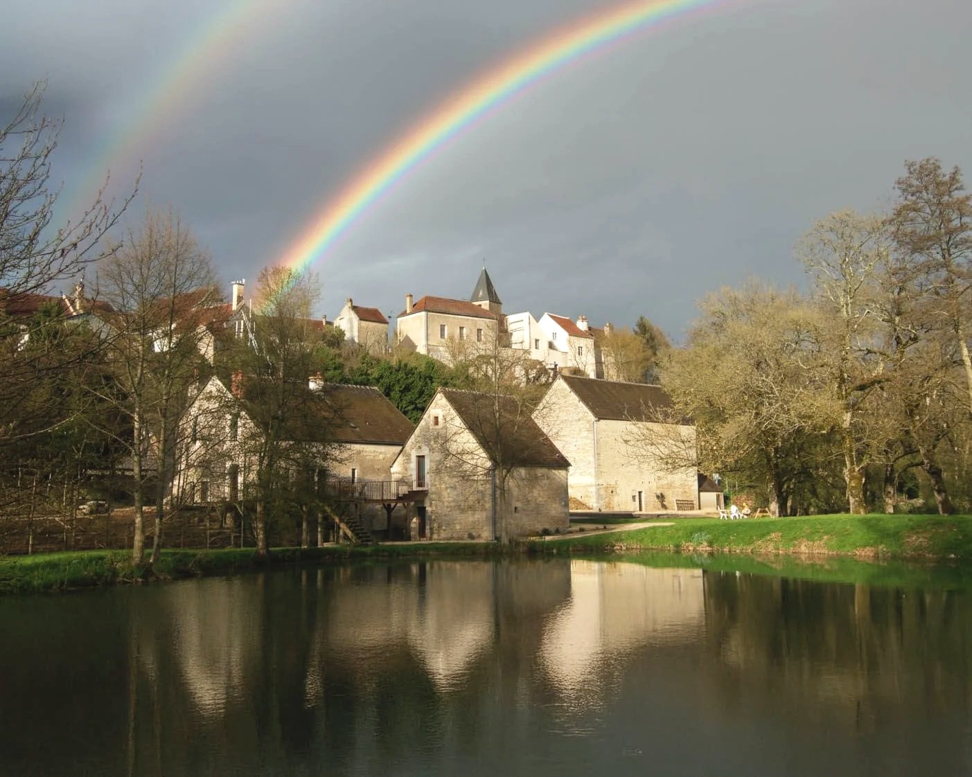 A vibrant double rainbow arches over a cluster of traditional stone buildings and a church in a French village, with their reflections shimmering in a calm mill pond in the foreground.