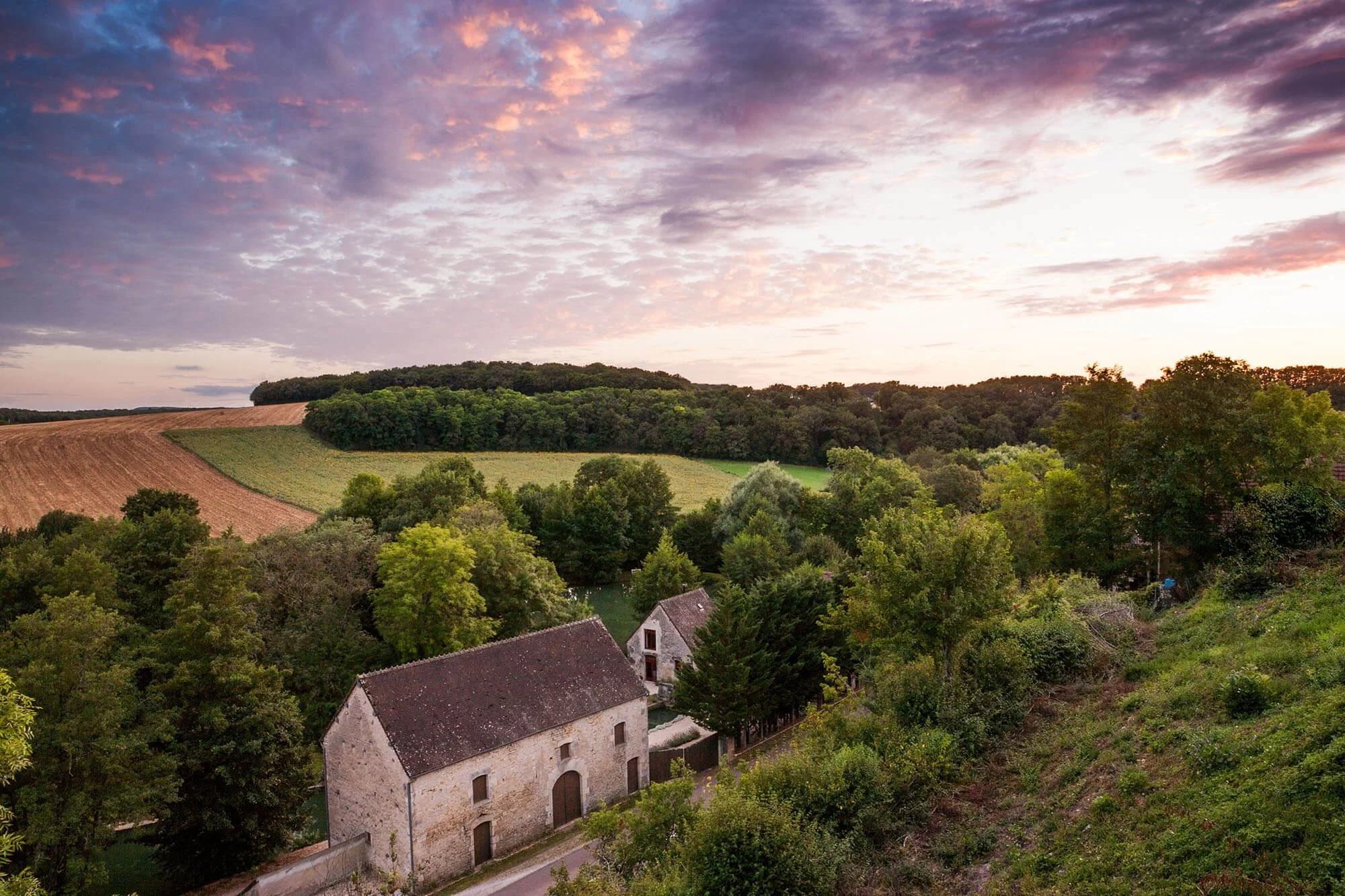 Elevated wide-angle view of the historic Rix Mill stone buildings in a lush Burgundy valley under a vibrant pink and purple sunset sky
