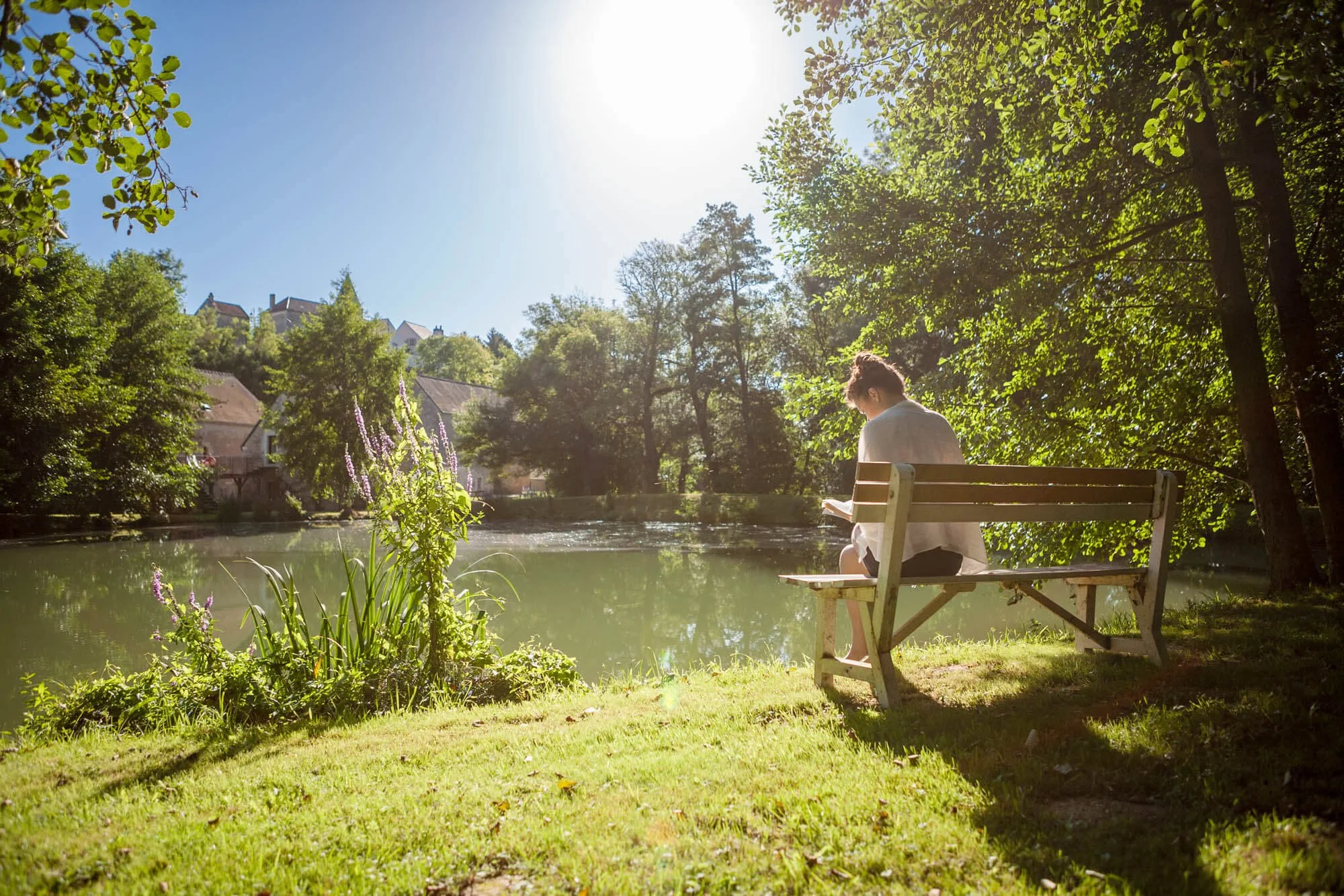 Woman sitting on a wooden bench by a quiet lake with Rix village on the hillside in the background