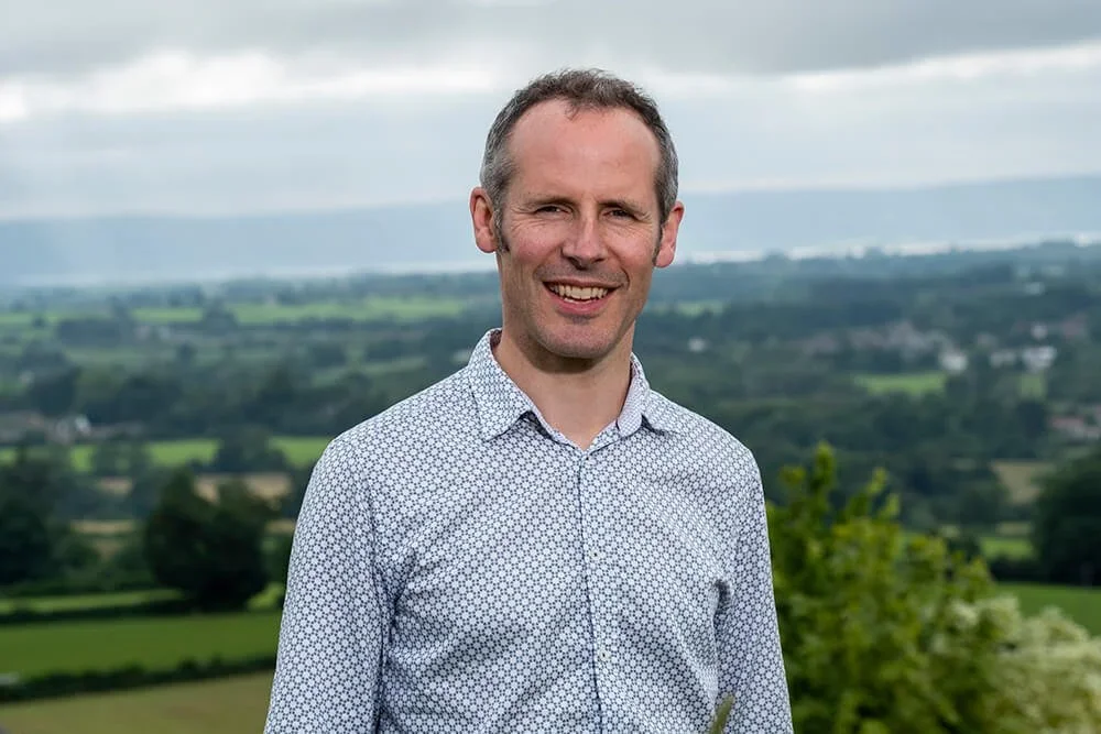 A smiling man standing outdoors in front of a green landscape with fields, trees, and distant mountains under an overcast sky.