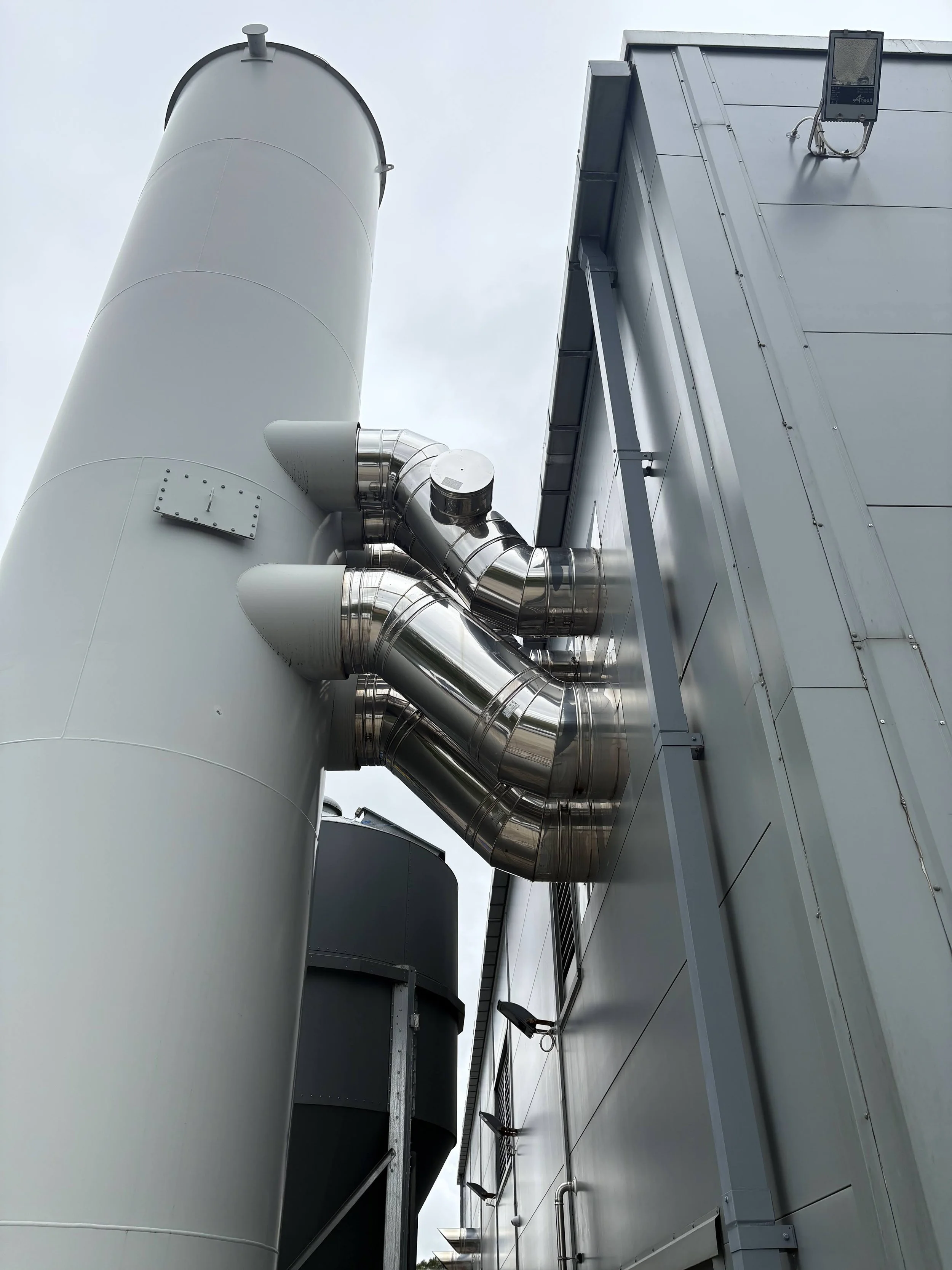 Close-up view of industrial building exterior with metal ducts, pipes, and a tall chimney against a cloudy sky.