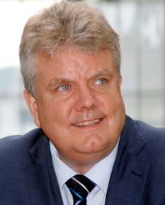 A middle-aged man with gray hair wearing a navy suit and striped tie, smiling, indoors with a large window behind him.