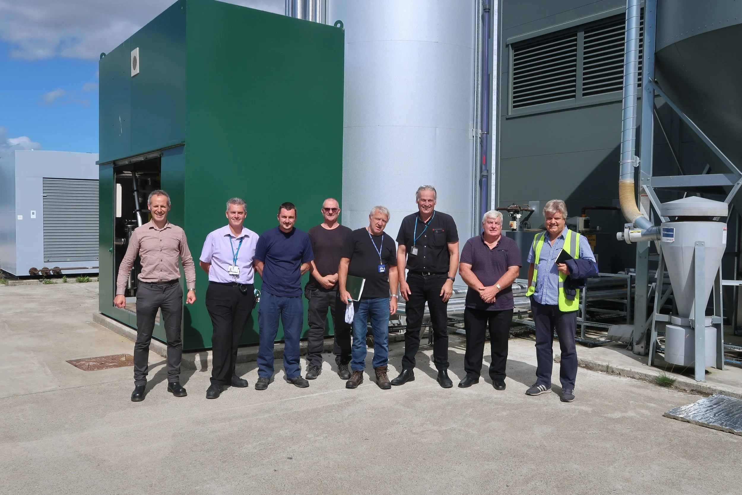A group of nine men standing outside in front of large industrial equipment, posing for a photo on a sunny day.
