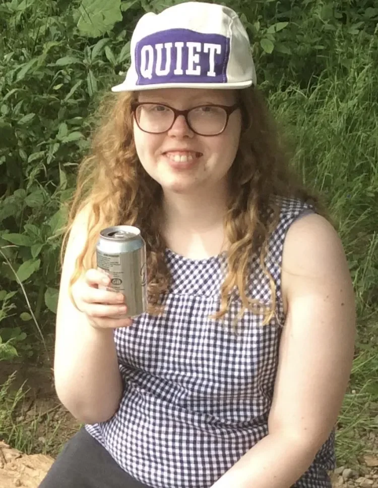 A woman with long curly hair and glasses, wearing a black and white checkered sleeveless top, holding a soda can, and sporting a white cap with a purple patch that says 'QUIET'. She is sitting outdoors with green foliage in the background.