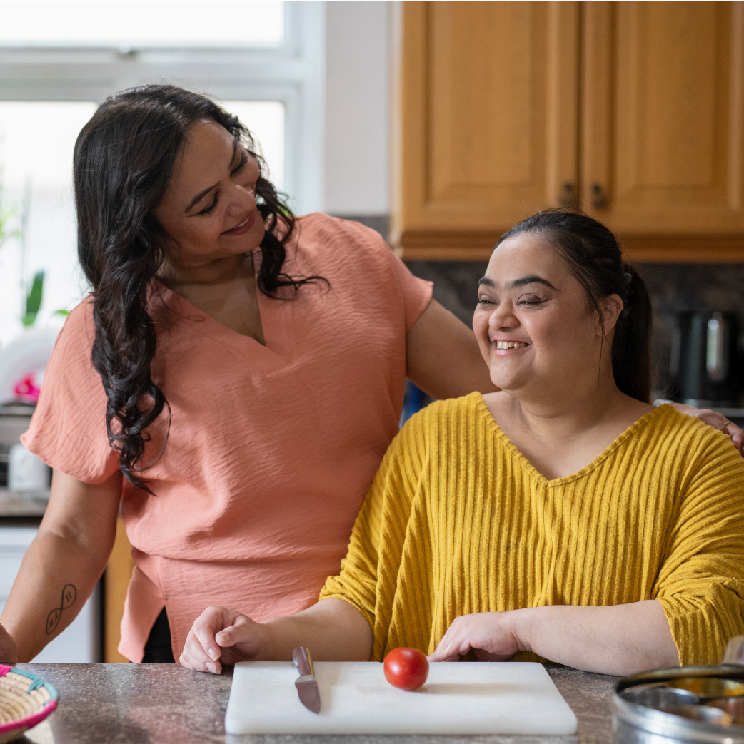 Two women in a kitchen smiling at each other; one woman is seated with a tomato and a knife in front of her, the other woman is standing beside her with her hand on her shoulder.