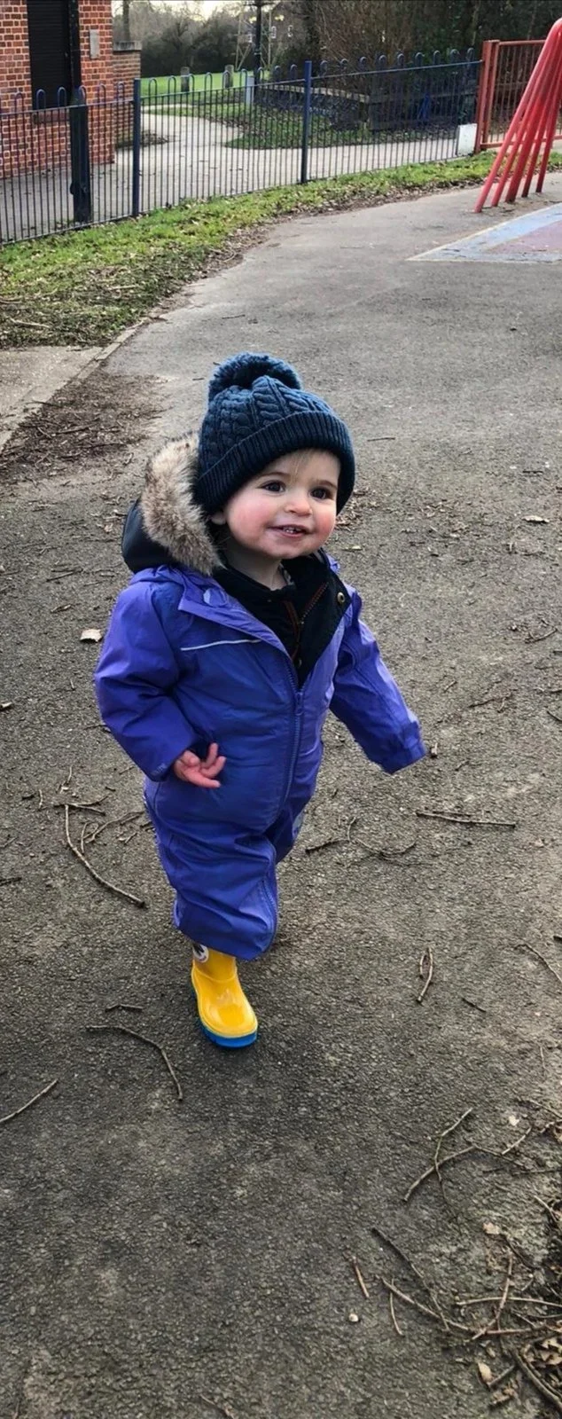 A young child dressed in a purple snowsuit, yellow rain boots, and a blue knit hat with a pom-pom, standing outdoors on a gravel surface near a playground.