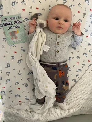 Baby lying on a sleep chart bed, holding a stuffed animal, dressed in pajamas with a playful print.