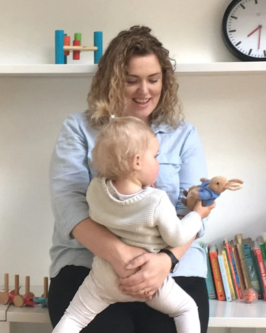 A woman with curly hair holding a young child with light hair, who is holding a stuffed animal rabbit. They are indoors, with a shelf of books and a clock in the background.