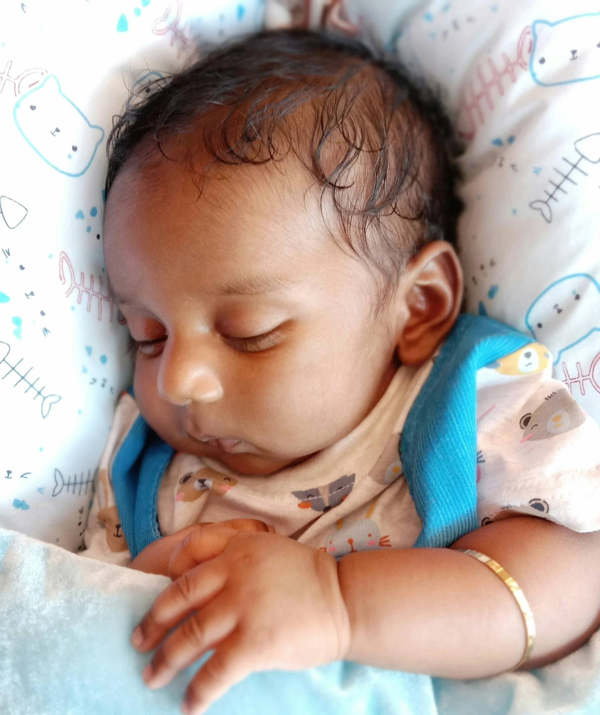 A sleeping baby with dark, slightly curly hair, lying on a white pillow with colorful animal prints, wearing a light-colored shirt with cute animal patterns and a blue strap, with a gold bracelet on the left wrist.