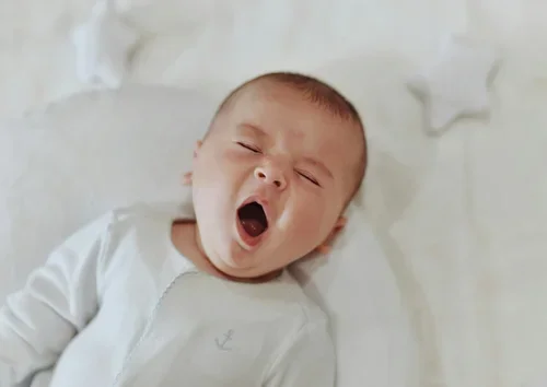 Young baby lying in bed, yawning with eyes closed and mouth wide open.