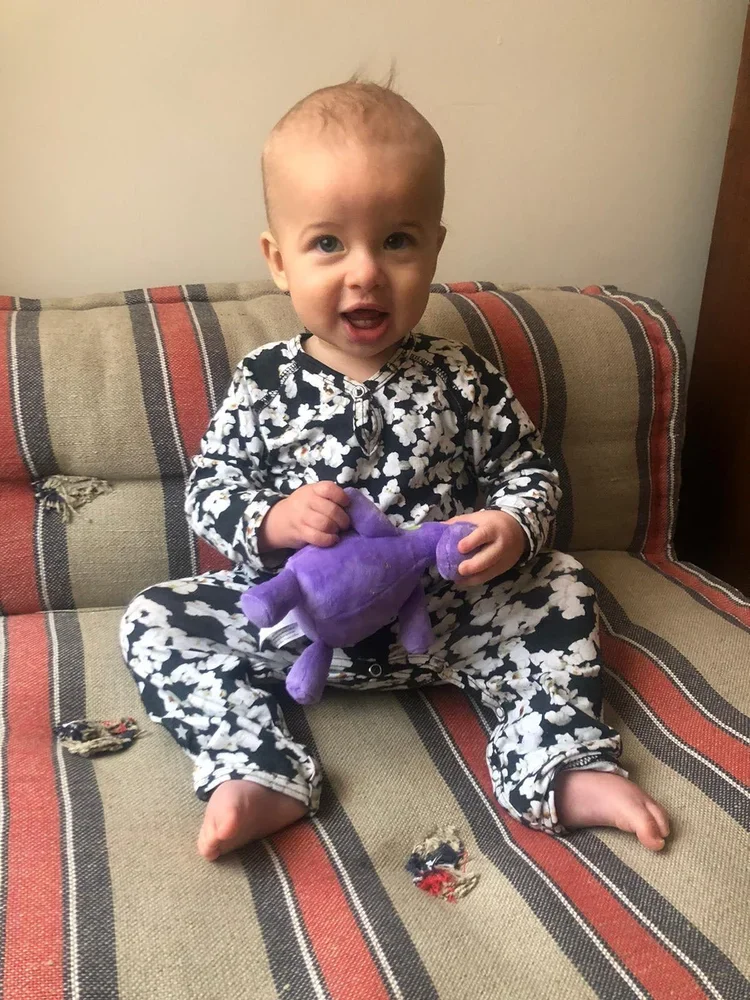 A baby sitting on a striped couch, holding a purple plush toy, wearing black and white pajamas with a floral pattern, with two torn spots on the couch fabric near the baby's feet.