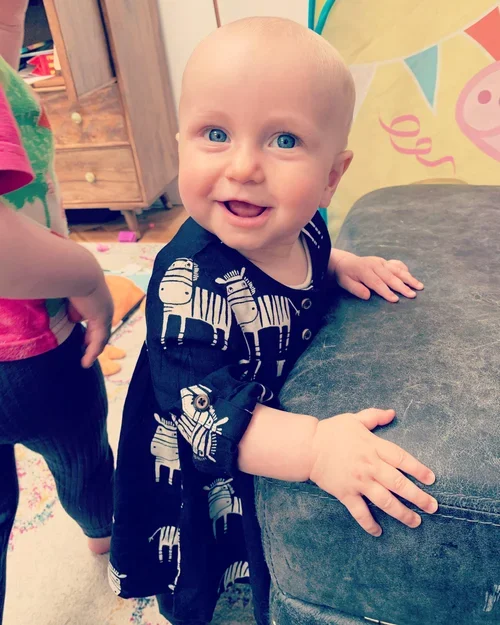 A smiling baby with blue eyes and light hair, standing and holding onto a piece of furniture in a colorful playroom.