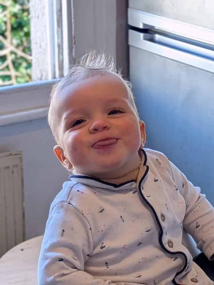 Happy young child with light blonde hair, sticking out tongue, wearing pajamas, sitting indoors near a window and a metallic silver refrigerator.