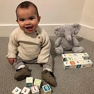 A happy baby sitting on the floor with toy blocks and a plush elephant.