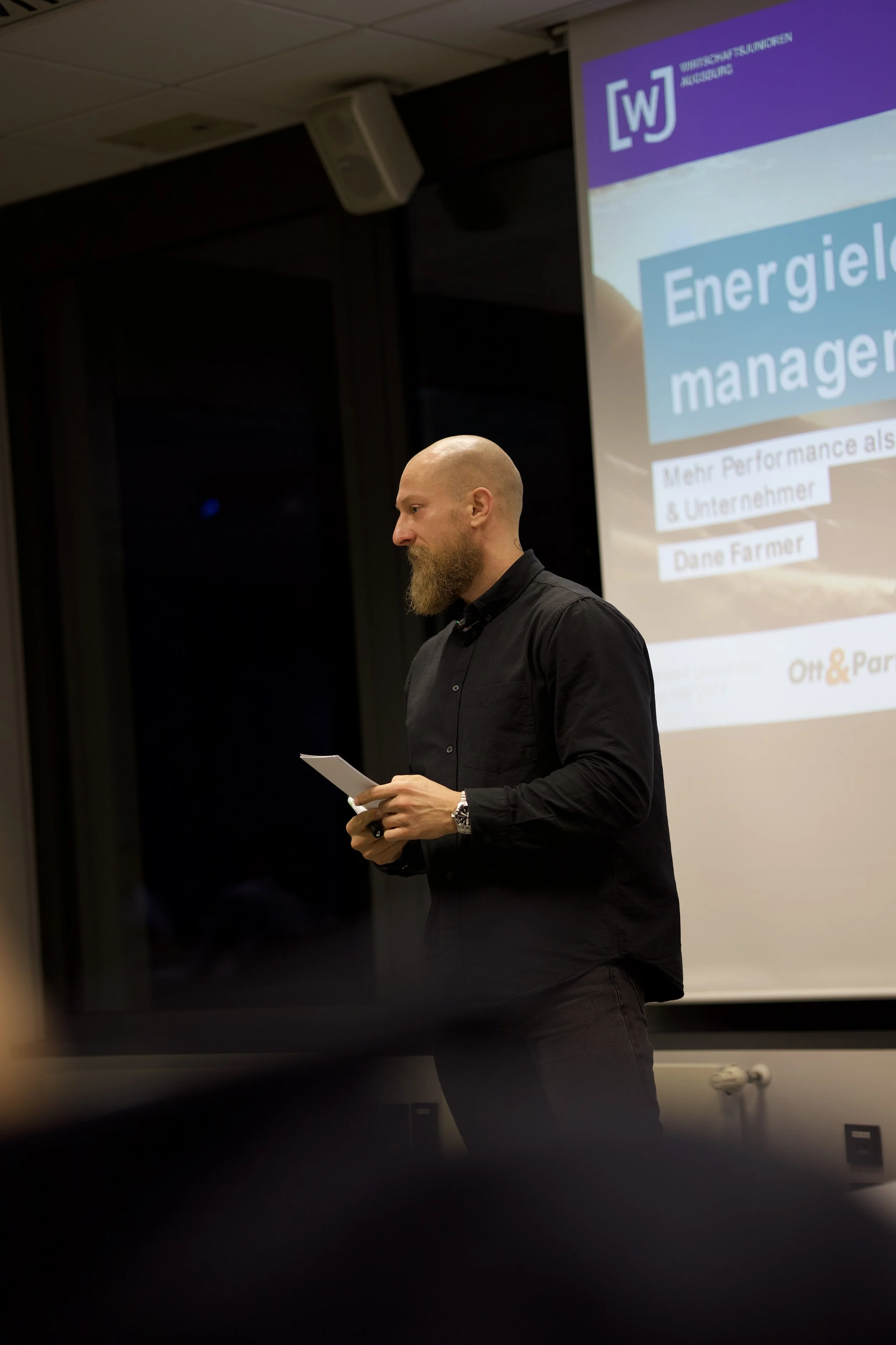 Man with a beard and glasses standing in front of a presentation screen, holding a notepad and pen, with a slide on the screen titled 'Energiel management'.