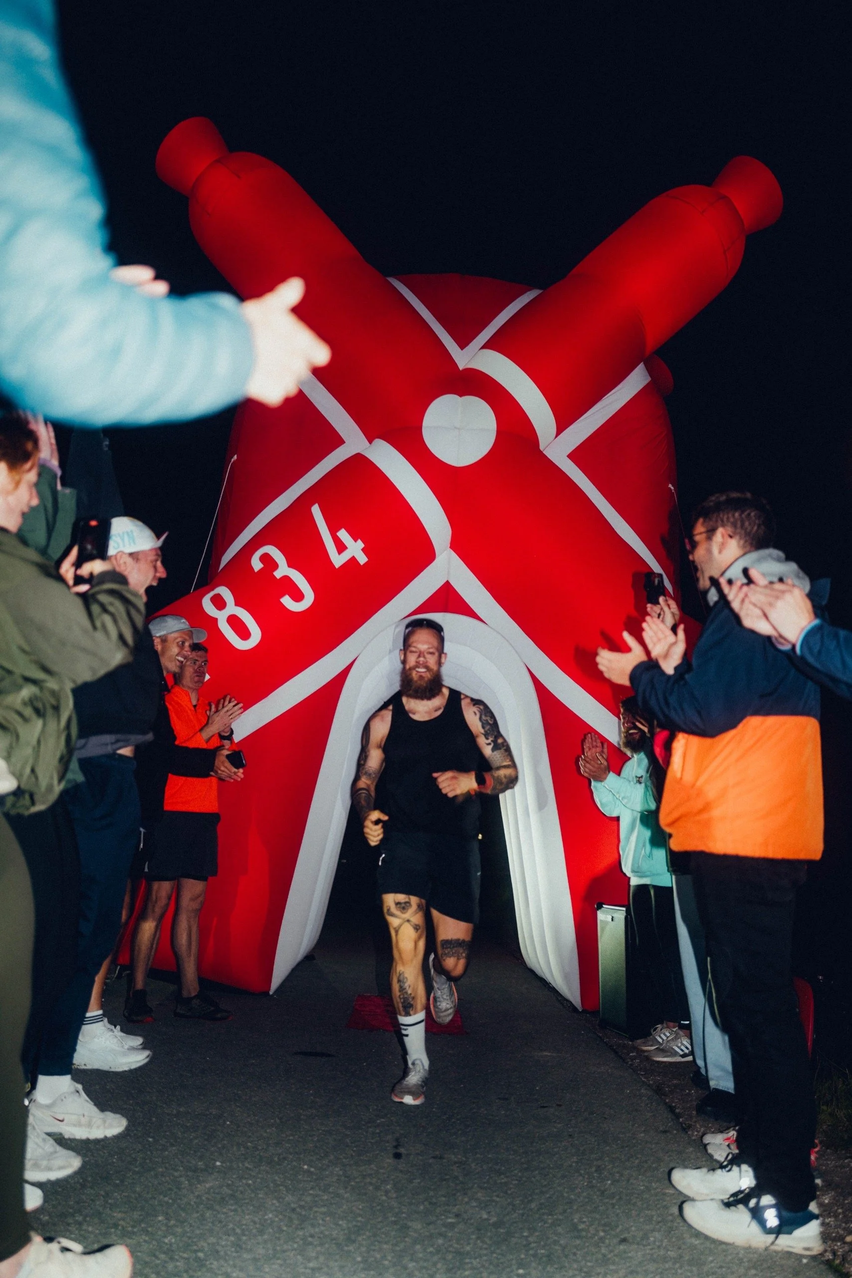 A man with tattoos running through an inflatable archway shaped like a red rocket ship at night, surrounded by cheering spectators.