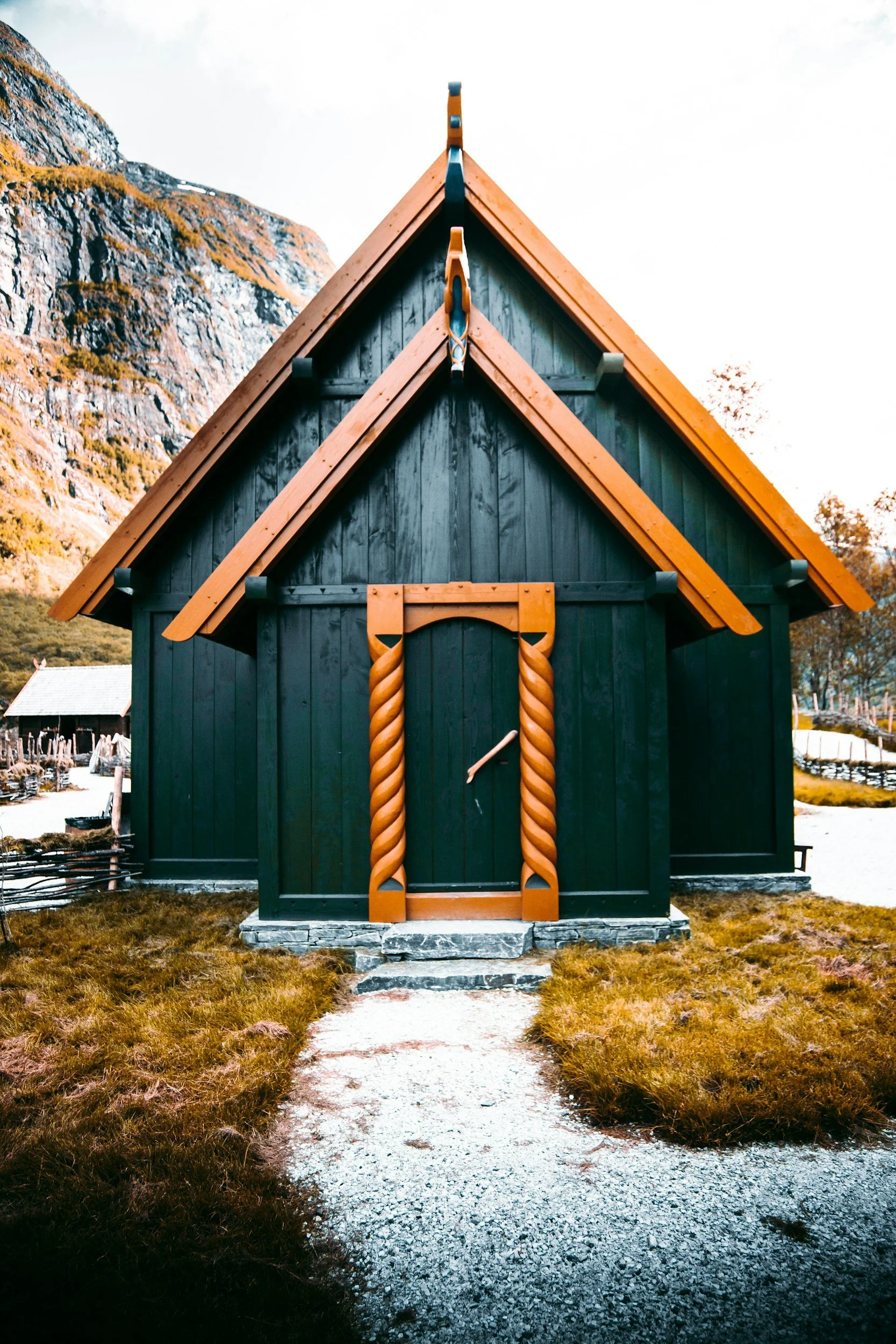 A small, black wooden house with ornate, twisted wooden door and matching twisted door frame. It has a steep, orange-colored roof and is situated outdoors with a mountain in the background and a gravel pathway leading to the entrance.