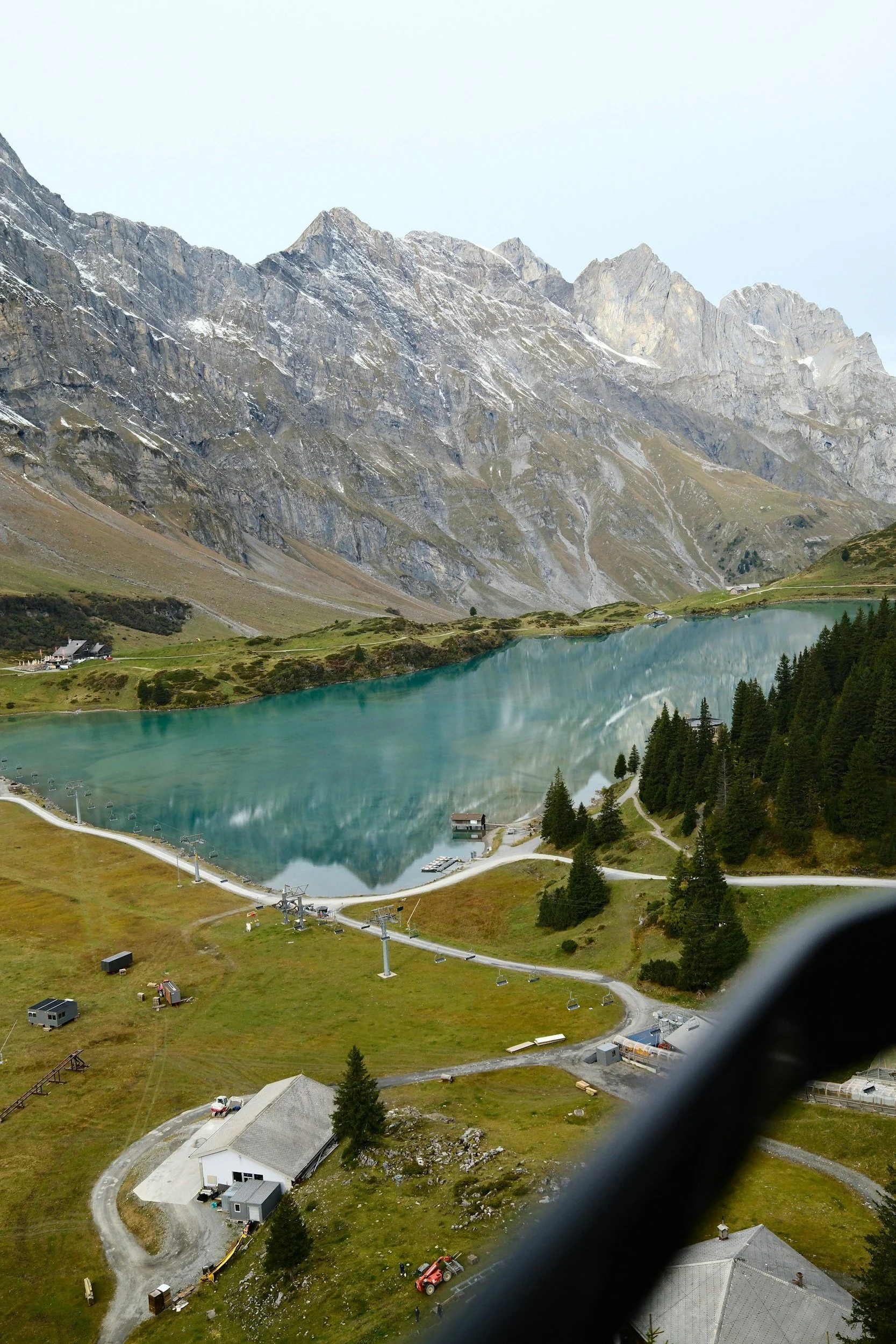 Scenic view of a mountain lake with clear blue water, surrounded by green grass, trees, and rocky mountain peaks in the background. A small building and chairlift cables are visible in the foreground.