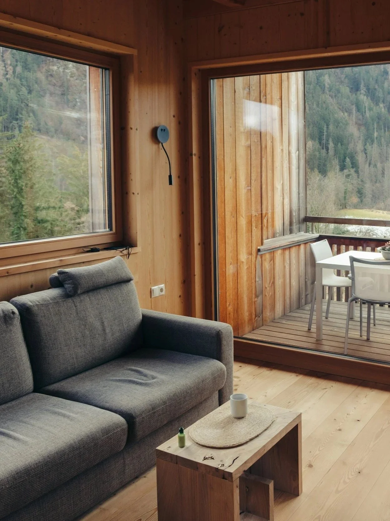Living room with gray sofa, small wooden coffee table with a white mug and small bottle, inside a wood-paneled house with large windows overlooking a forested landscape and a balcony with a table and chairs.