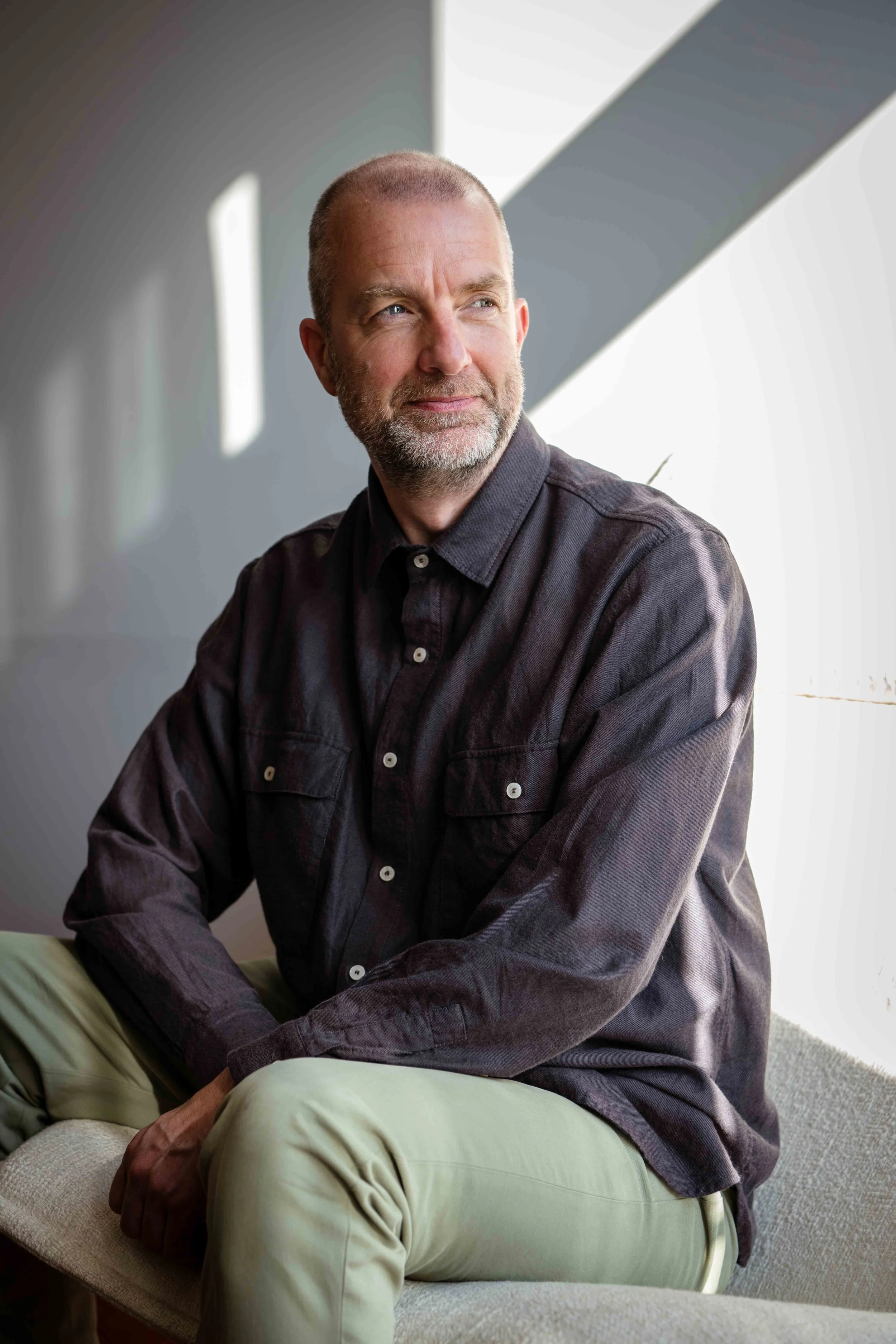 A middle-aged man with a beard and short hair sitting on a sofa near a window, wearing a dark shirt and light-colored pants, looking thoughtfully outside.