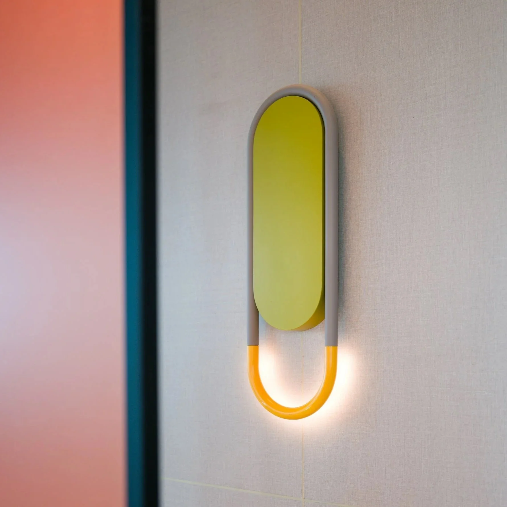 A modern wall-mounted light fixture with a green oval panel and an orange U-shaped light on a plain wall.