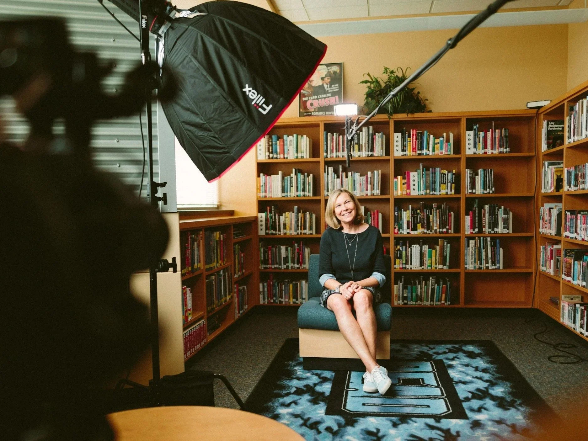 A woman sitting on a chair in a library, getting filmed with a professional lighting setup, with bookshelves filled with books behind her displaying Polaar Media's Video creation capability.
