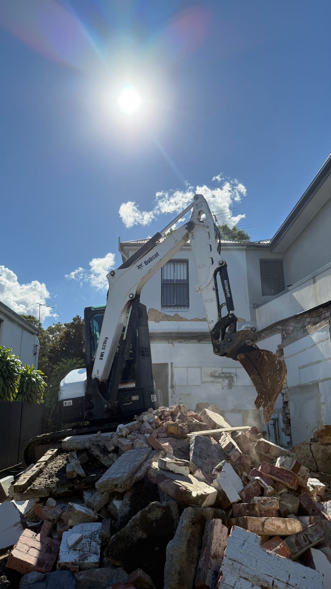 A small excavator tearing down a building wall, with rubble and debris on the ground, under a bright sunny sky.