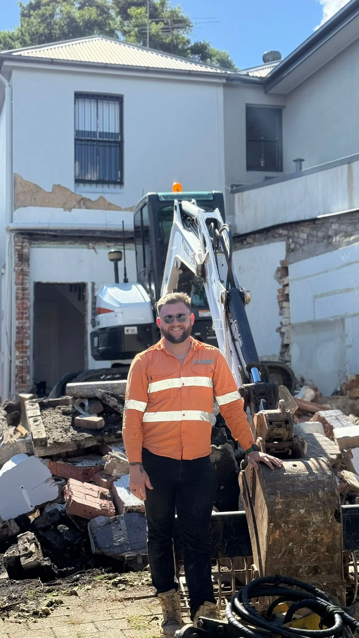 A man in an orange safety jacket and glasses standing in front of broken brick and concrete debris on a construction site with an excavator behind him.