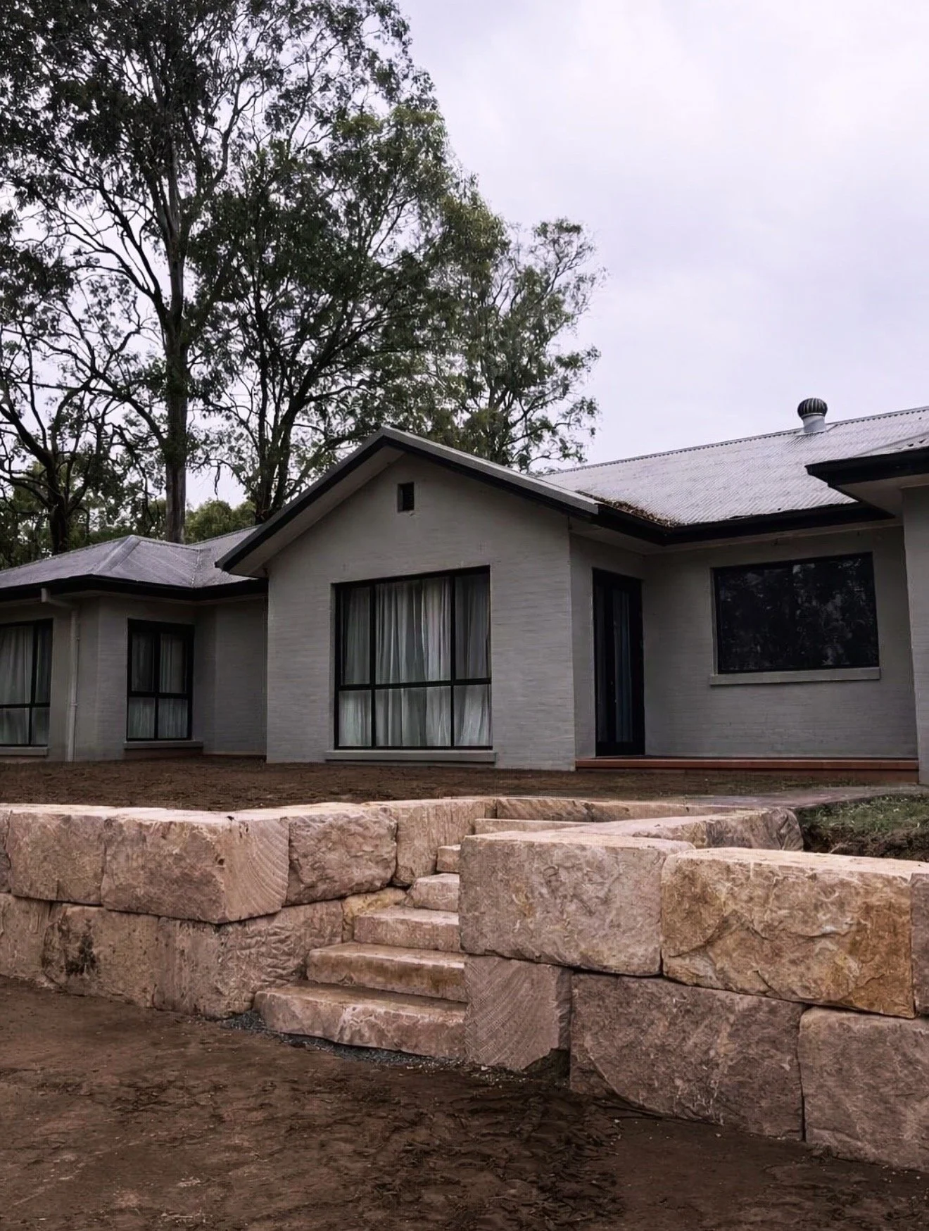 A house with dark window frames and a metal roof, surrounded by tall trees, with a stone staircase and retaining wall in the foreground.