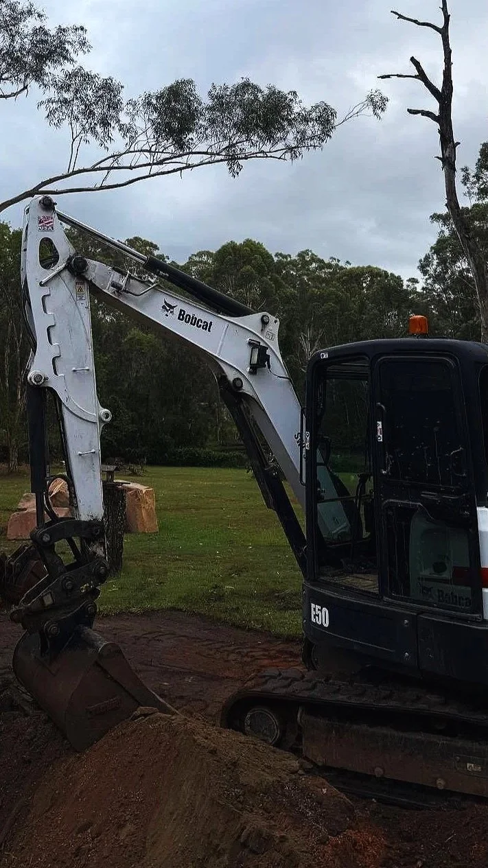 A Bobcat mini excavator moving dirt in a grassy outdoor area with trees in the background.