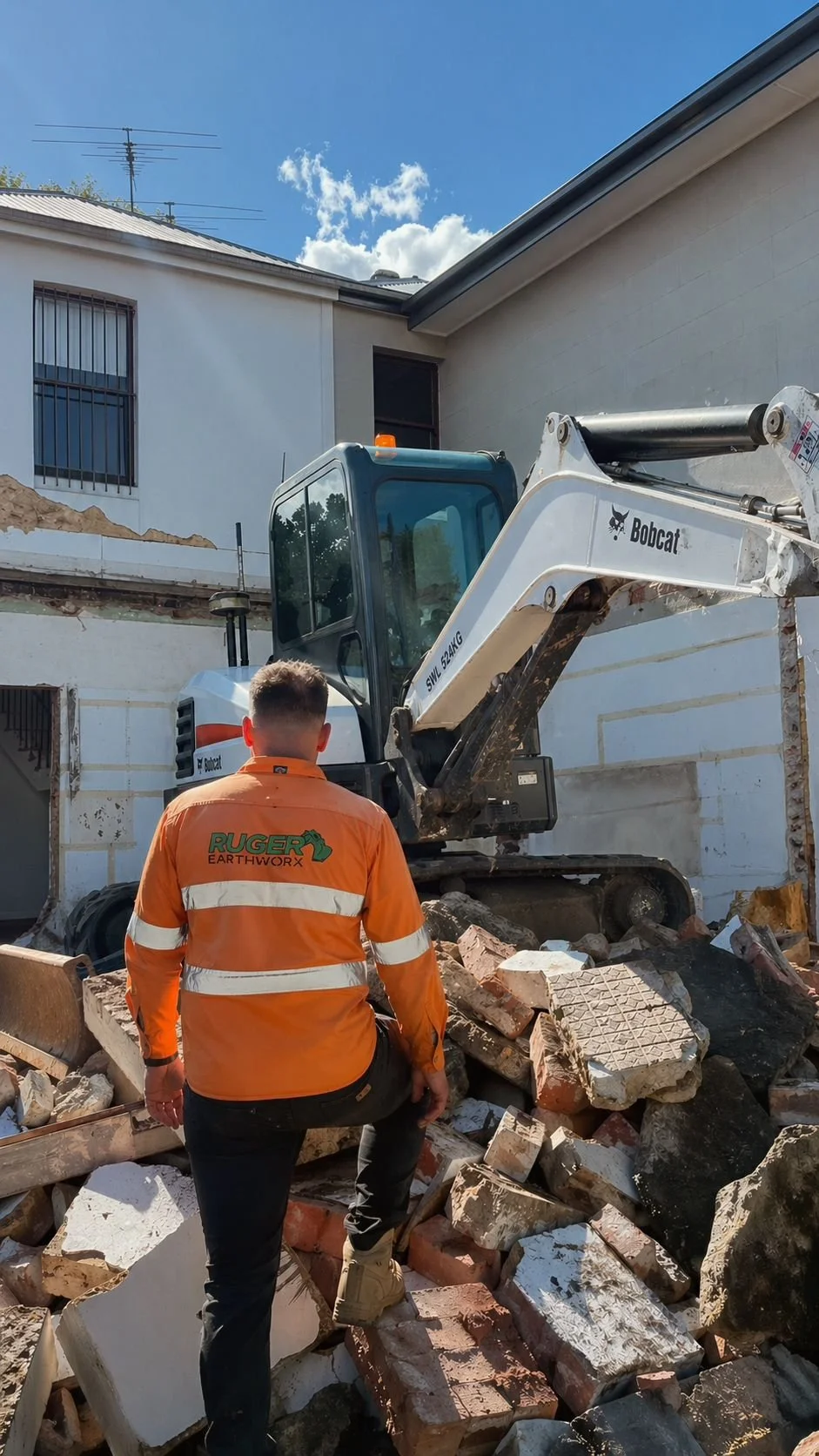 A construction worker wearing an orange jacket labeled 'Ruger EarthworX' watches as a small excavator demolishes a brick wall amidst debris, with a clear blue sky overhead.