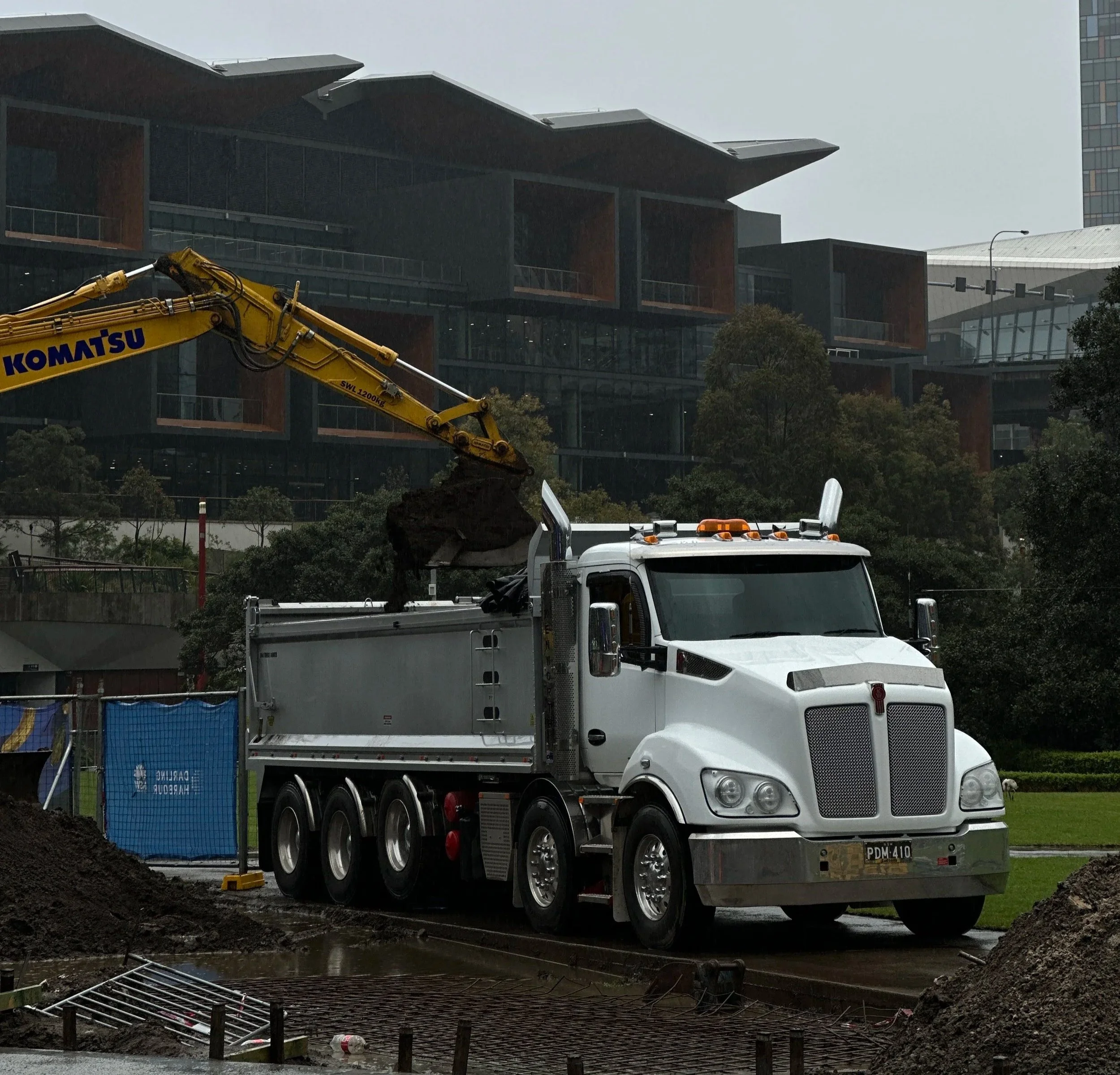 Construction site with a white truck and yellow Komatsu excavator in front of a modern building under construction.