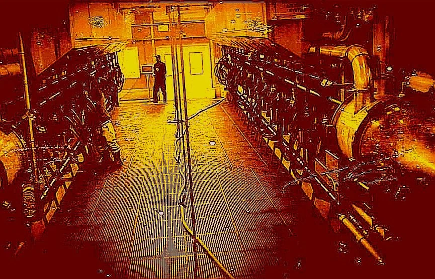 Inside a large industrial or mechanical room with metal walkways, large pipes, and equipment, illuminated with orange lighting, with a person standing at the far end.