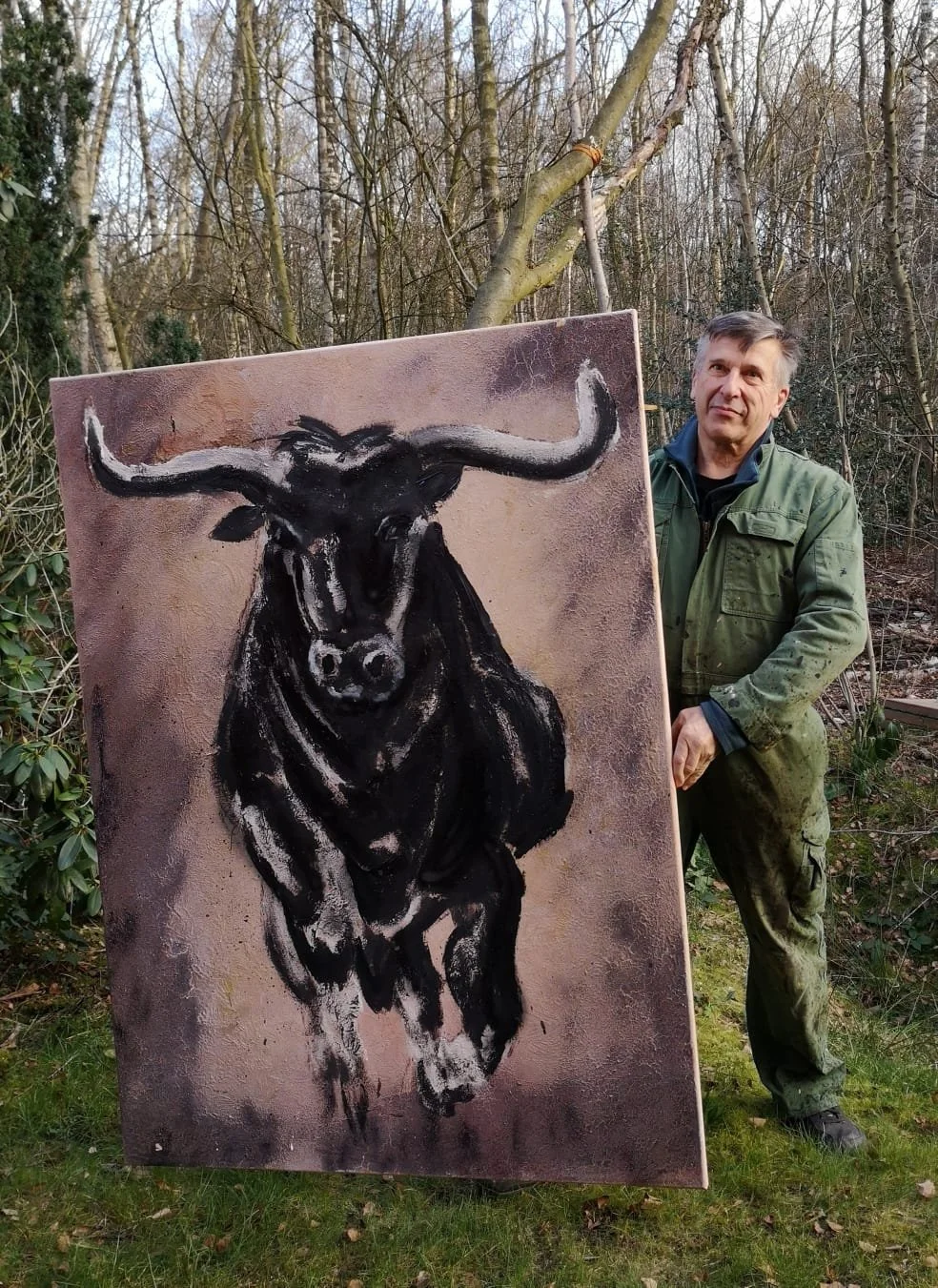 A man wearing a green jacket stands outdoors in a wooded area, holding a large canvas painting of a black bull with white horns, depicted in an expressive, abstract style.