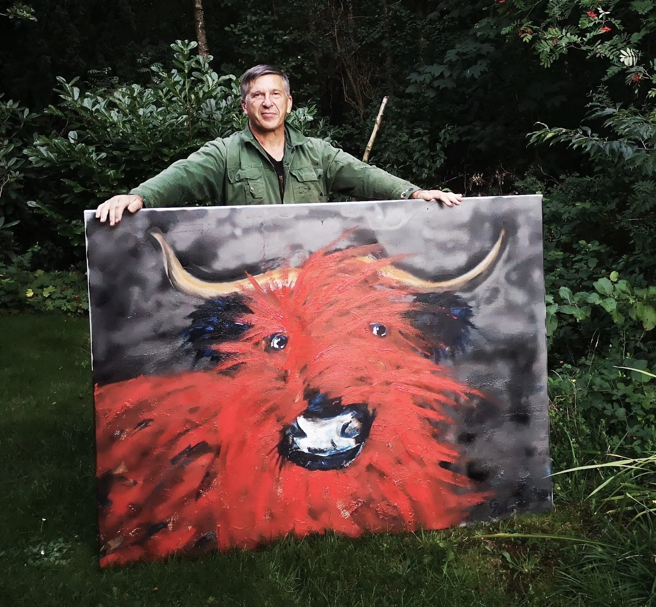 A man standing outdoors holding a large painting of a red furry animal with horns and big eyes, against a backdrop of green shrubbery and trees.
